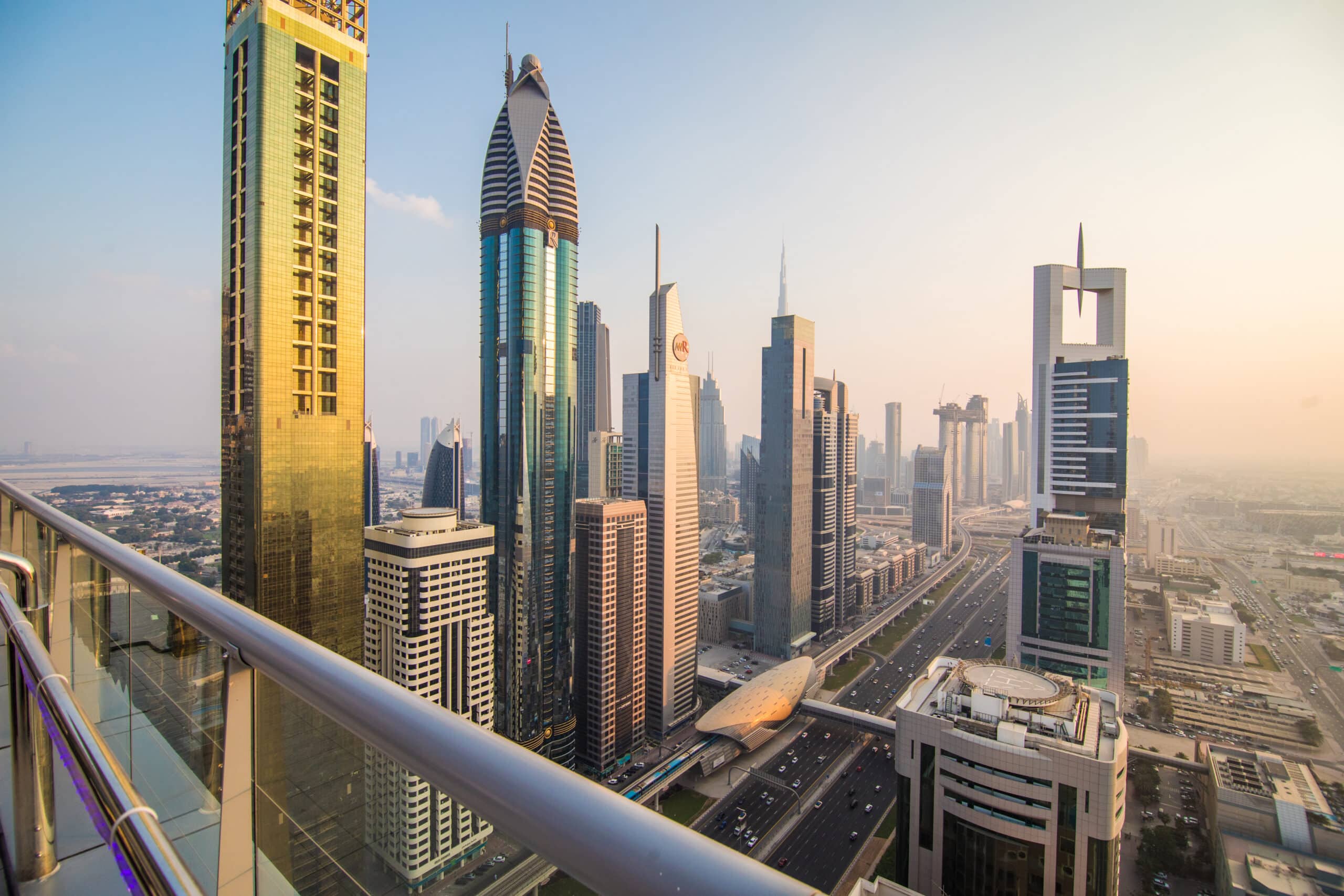 Dubai skyline at sunset with the Burj Khalifa illuminated against a vibrant orange and purple sky, surrounded by modern skyscrapers and intricate highways glowing with city lights.